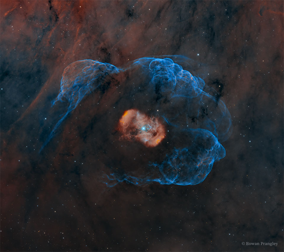 A blue star is seen in the center of a red nebula 
itself surrounded by a faint blue nebula. The surrounding
starfield itself has a faint red-brown emission clouds.
Please see the explanation for more detailed information.