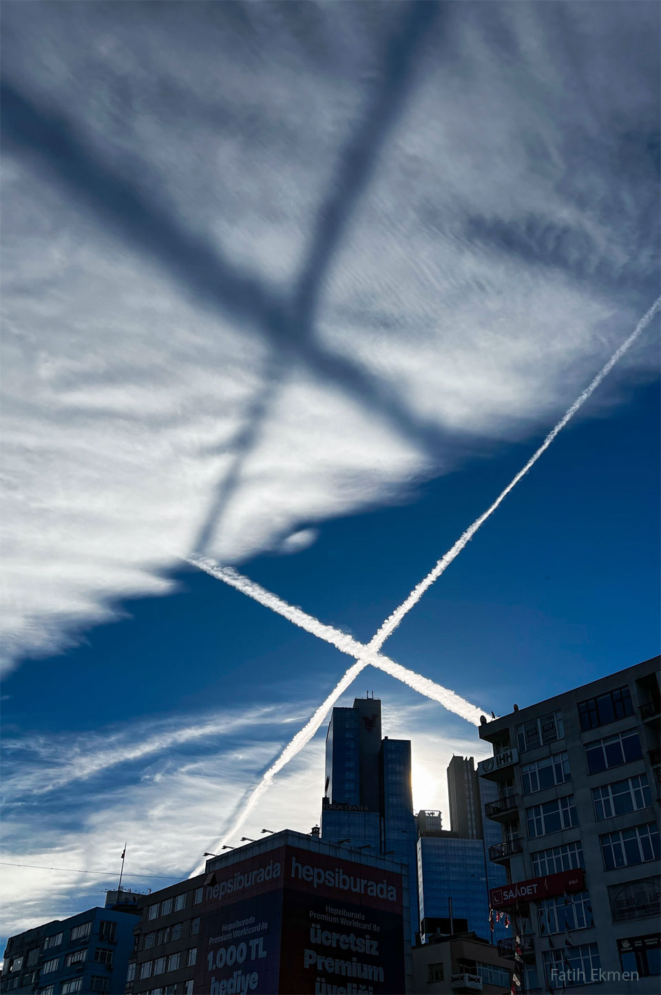Two airplane contrails, crossing in an X, are shown across
the middle of the image. They are bright white against a dark
blue background. A high cloud deck is seen above the crossing,
sunlit, contrails. A low Sun creates a dark shadow X on the high
while clouds. A row of buildings runs across the lower
part of the image. 
Please see the explanation for more detailed information.